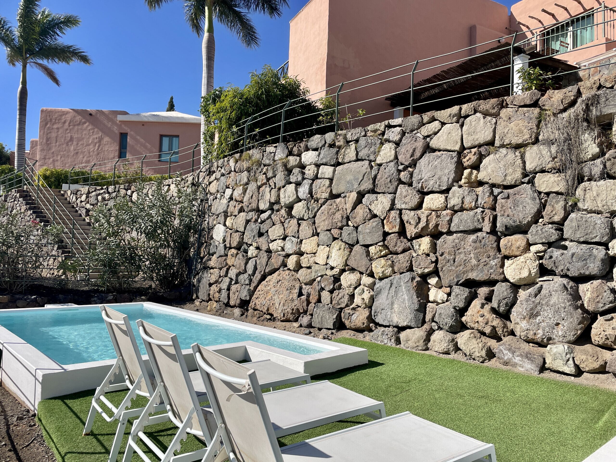 En swimmingpool med solstolar, en stenmur och en trappa som leder upp till en byggnad.