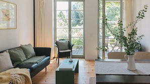 A living room with a sofa, a chair, a coffee table, and a dining table. There are large windows and a view of greenery outside.