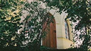A church with a prominent wooden door and arched windows, surrounded by dense foliage.