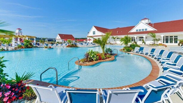 Una piscina con sillas de descanso, una pequeña isla con plantas y un edificio con techo rojo.
