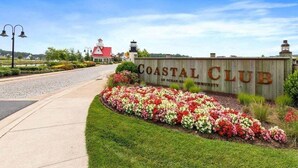 Señal del Coastal Club con un parterre floral, un edificio de techo rojo y un faro al fondo.