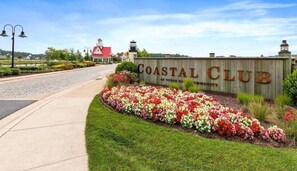 A Coastal Club sign with a landscaped flower bed, a red-roofed building, and a lighthouse in the background.