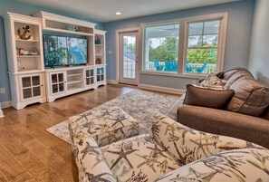 A living room with a large flat-screen TV, white entertainment center, brown sofa, and a view of the outdoors through a large window. - Bayside * 5BR New Luxury House (Selbyville)