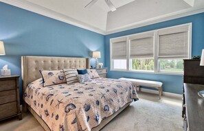 A bedroom with a tufted headboard, a large bed with a patterned bedspread, a bench, a dresser, and a window with blinds. - Coastal Club * 5BR Luxury Beach House (Lewes)