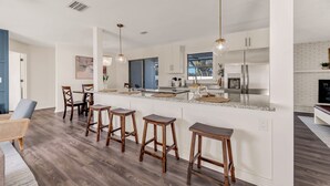 Una cocina moderna con muebles blancos, un refrigerador de acero inoxidable y una encimera de mármol. Hay cuatro taburetes de madera en la isla.