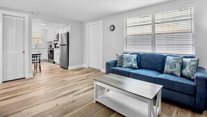 Uma sala de estar moderna com um sofá azul, piso de madeira, uma mesa de centro e vista para a cozinha ao fundo.