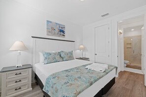 A bedroom with a bed, two bedside tables, a nightstand, a painting, and a bathroom with a shower.