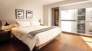 A modern bedroom with a large bed, wooden flooring, and a large window with blinds.