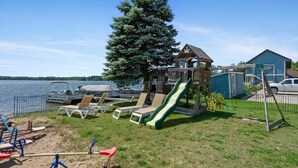 A lakeside playground with a slide, swings, and a small house structure.