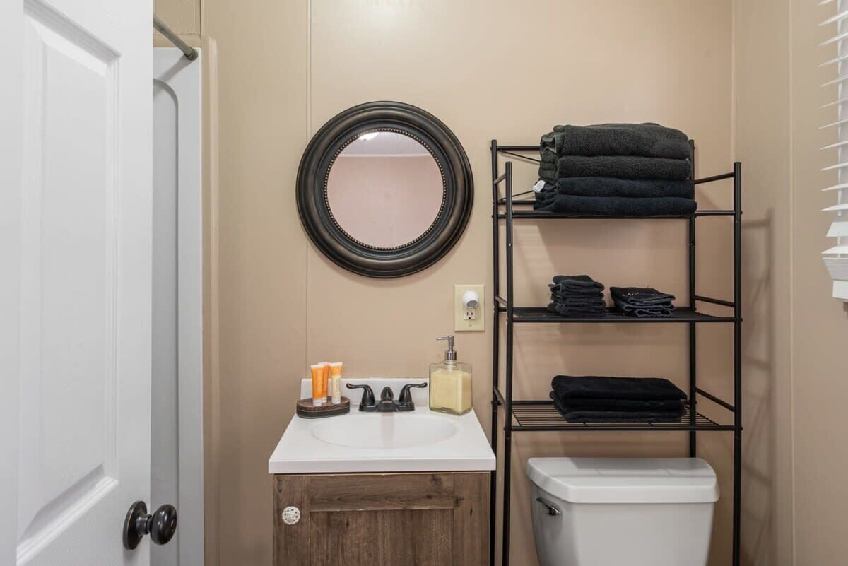 Een badkamer met een wit wastafelmeubel, een ronde spiegel, een plank met opgevouwen handdoeken, toiletartikelen en een wit toilet.