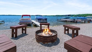 A lakeside deck with a fire pit, wooden benches, and boats docked at the pier.