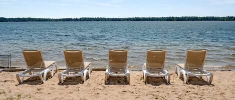 A row of beach chairs facing a calm lake with a forested shoreline.