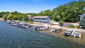 A marina with several boats docked, a blue building with a porch, and a forested area in the background.