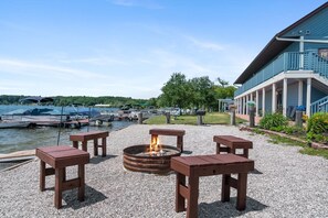 A waterfront patio with a fire pit, wooden benches, and a building with a covered walkway.