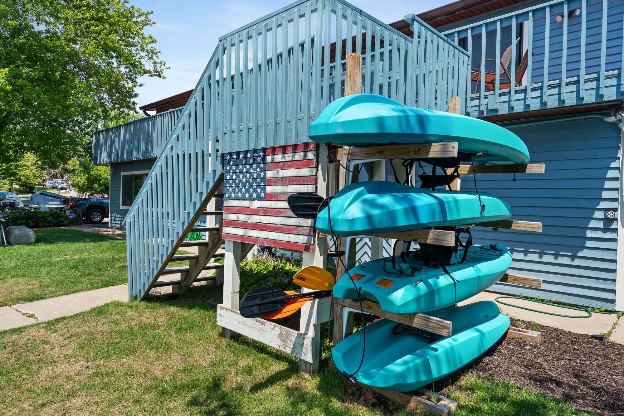 A house with a blue exterior and a flagpole with an American flag. There are four kayaks stacked on a wooden structure in front of the house.
