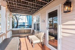 A covered porch with a cushioned seating area, a wooden deck, and a view of a blue house in the background.