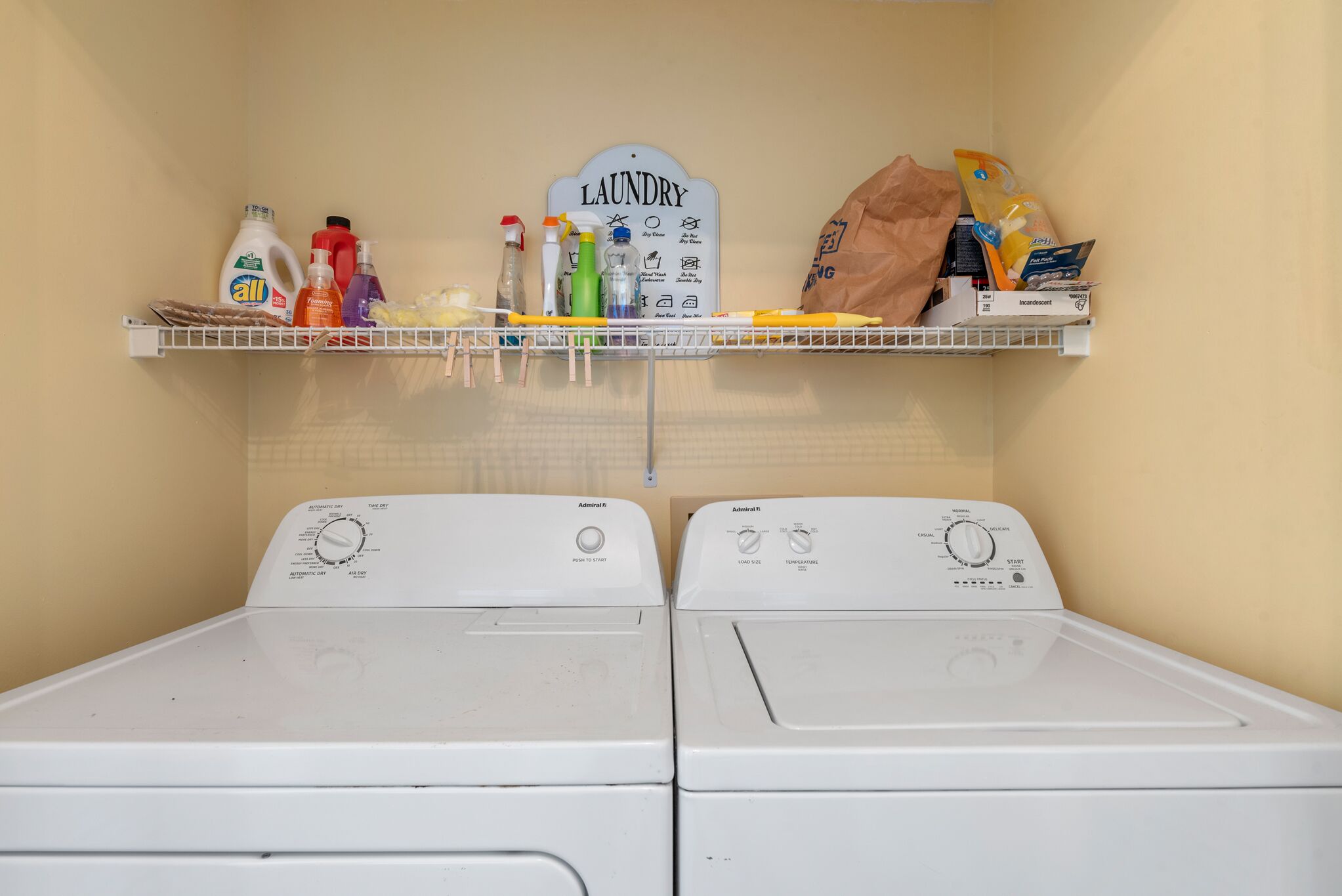 A laundry area with two washing machines, a shelf with various laundry supplies, and a laundry sign.