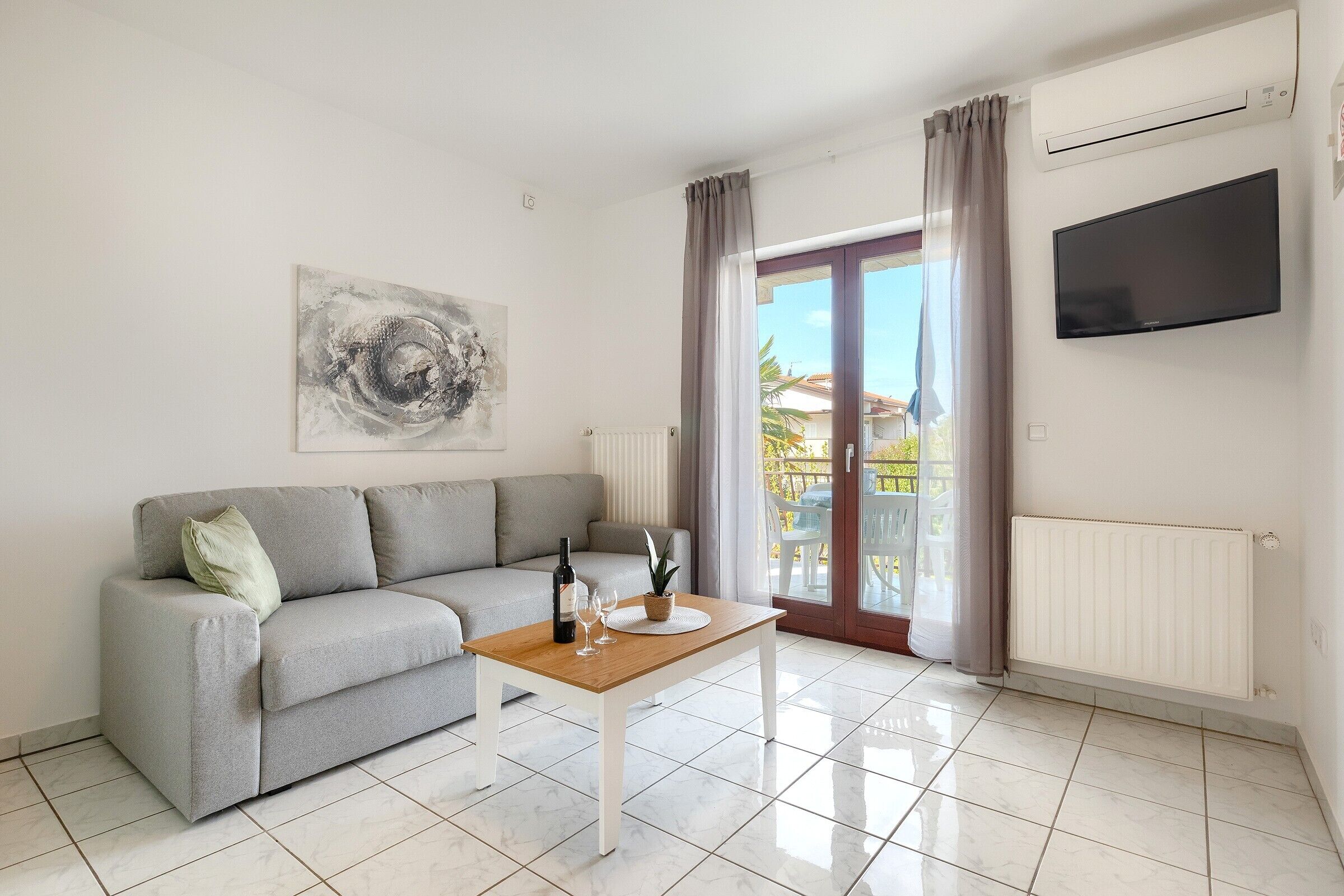 A modern living room with a grey sofa, a wooden coffee table, a TV mounted on the wall, and a sliding glass door leading to an outdoor area.