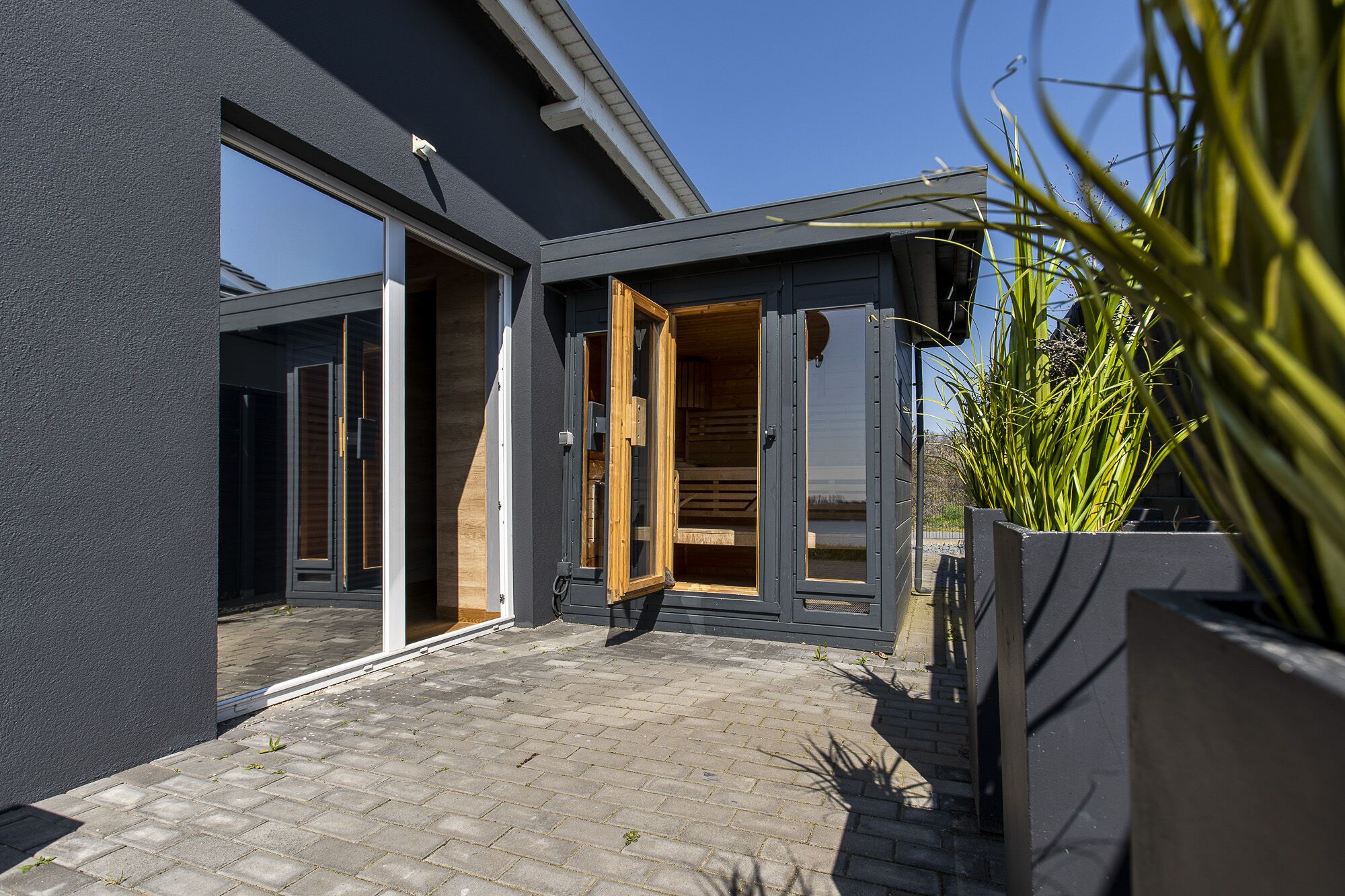 Una casa moderna con camino de entrada pavimentado, amplias puertas de vidrio y un interior de madera visible a través de la puerta abierta.