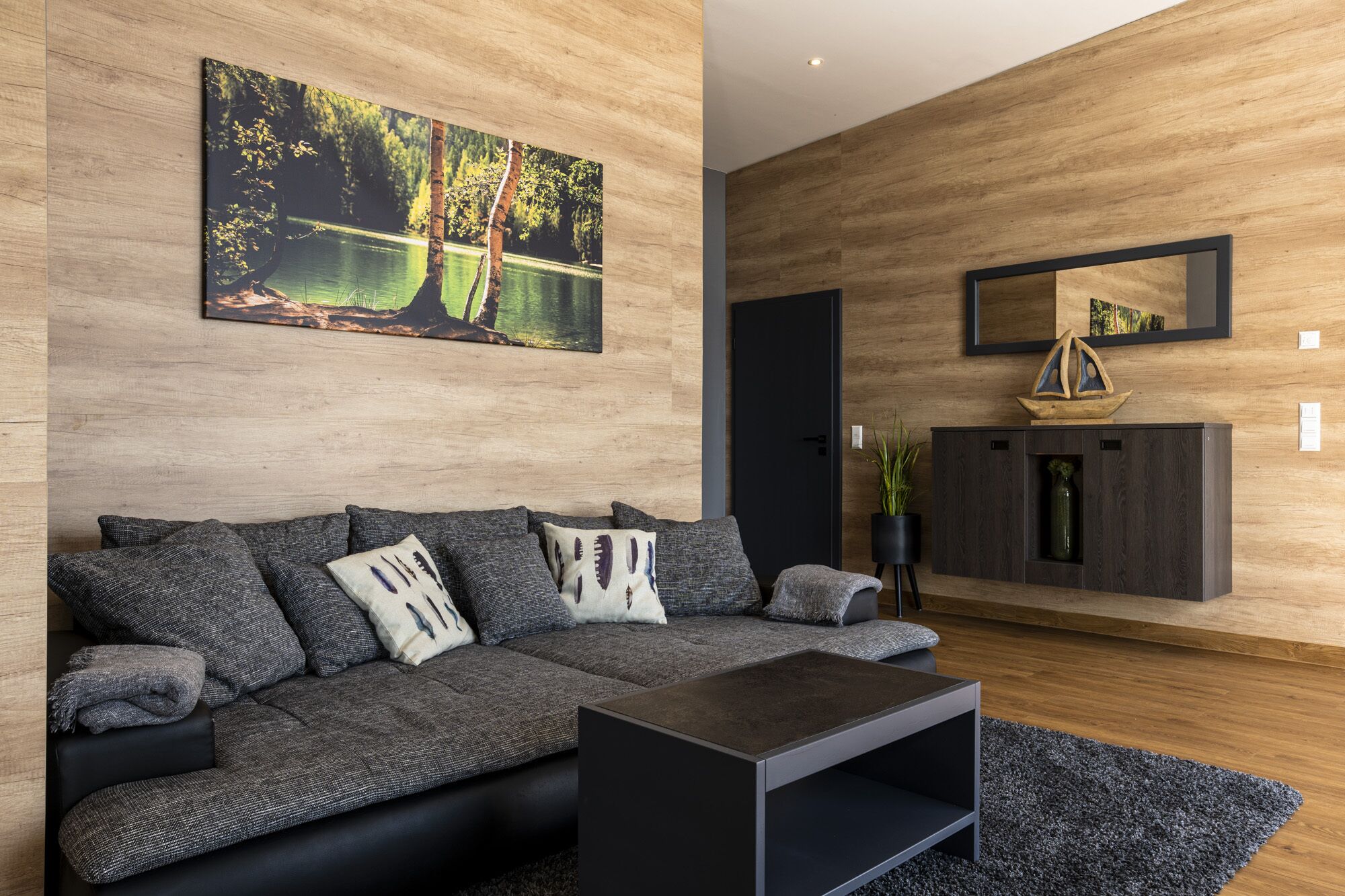 A modern living room with a grey sofa, a coffee table, and a wooden wall with a painting of a lake.