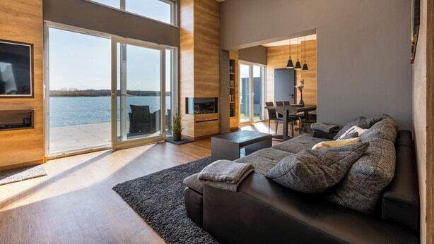 Ein modernes Wohnzimmer mit einem großen Schiebeglasfenster, das auf einen Balkon mit Blick auf ein Gewässer führt.