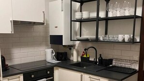 Uma cozinha compacta com armários brancos, bancada preta, forno embutido e prateleira com pratos e copos.