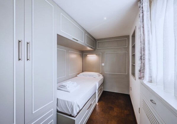 A small, narrow room with a single bed, built-in wardrobe, and a window with curtains.