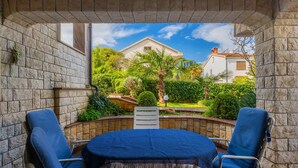 A covered patio with a table and two chairs, a stone wall, and a view of a house and greenery. - Beautiful spacious apartment (No. 5) with 2 bedrooms 300m from the beach (Porec)