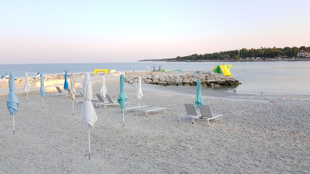 A beach with rows of sun loungers and umbrellas, a rocky breakwater, and a calm sea. - Beautiful spacious apartment (No. 5) with 2 bedrooms 300m from the beach (Porec)