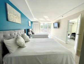 Two large beds with white linens and grey headboards, a flat-screen TV, and a large window offering a view of the outdoors.
