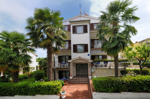 A multi-story building with a central entrance, surrounded by lush greenery and palm trees. - Beautiful spacious apartment (No. 2) with 2 bedrooms 300m from the beach (Porec)