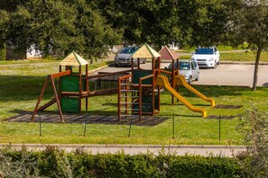 A playground with climbing structures and slides, surrounded by greenery and parked cars.