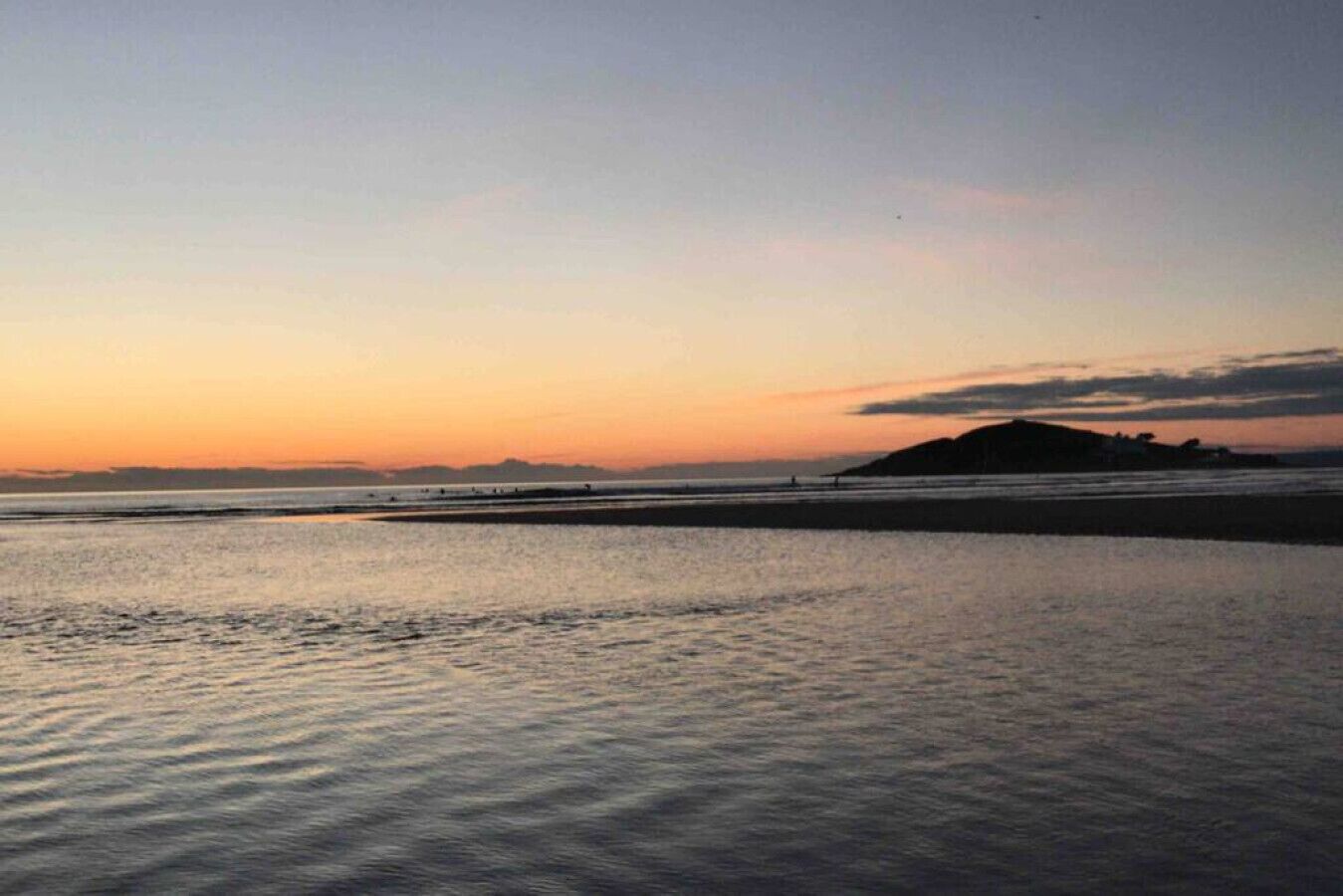 Une plage au coucher du soleil, avec une eau calme et des reliefs lointains.