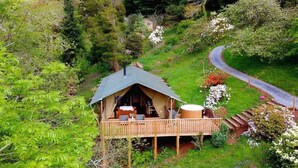Una casa en el árbol con terraza de madera y bañera de hidromasaje, rodeada de exuberante vegetación y un sendero sinuoso.