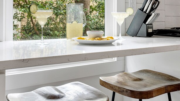 A kitchen counter with a pitcher, a glass of a clear drink, and a plate of food. There are two stools in front of the counter. - Prime Location in Park Circle | The Aloha Cottage (North Charleston)