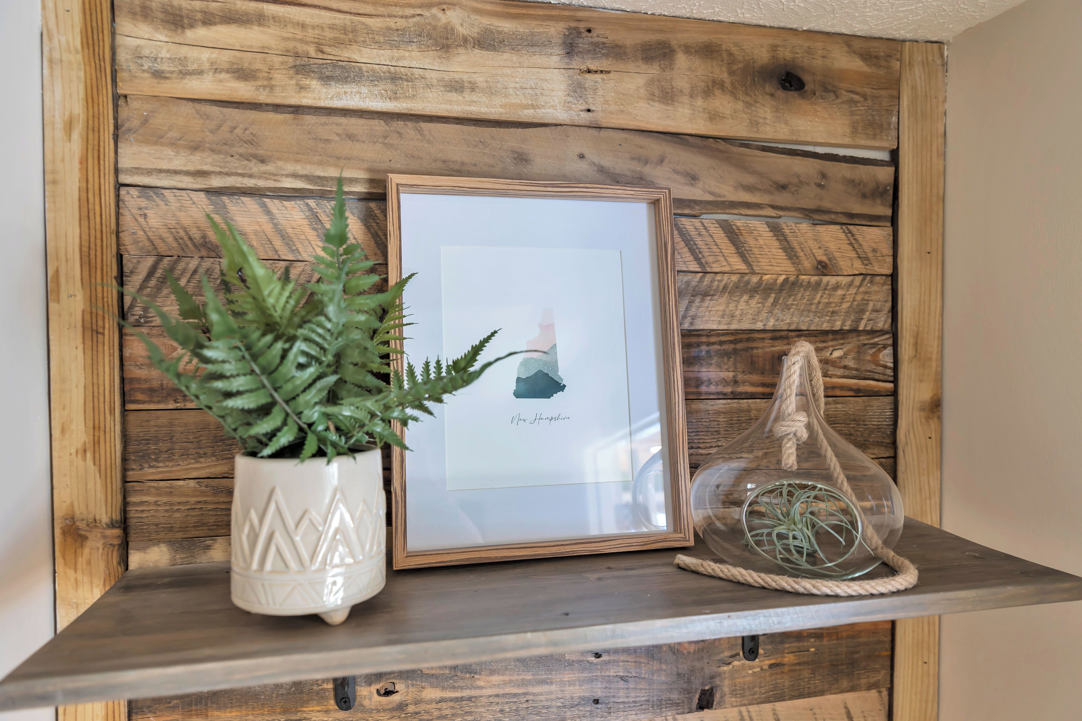 Une étagère en bois avec un tableau encadré, une plante en pot et un vase en verre orné d’une corde.