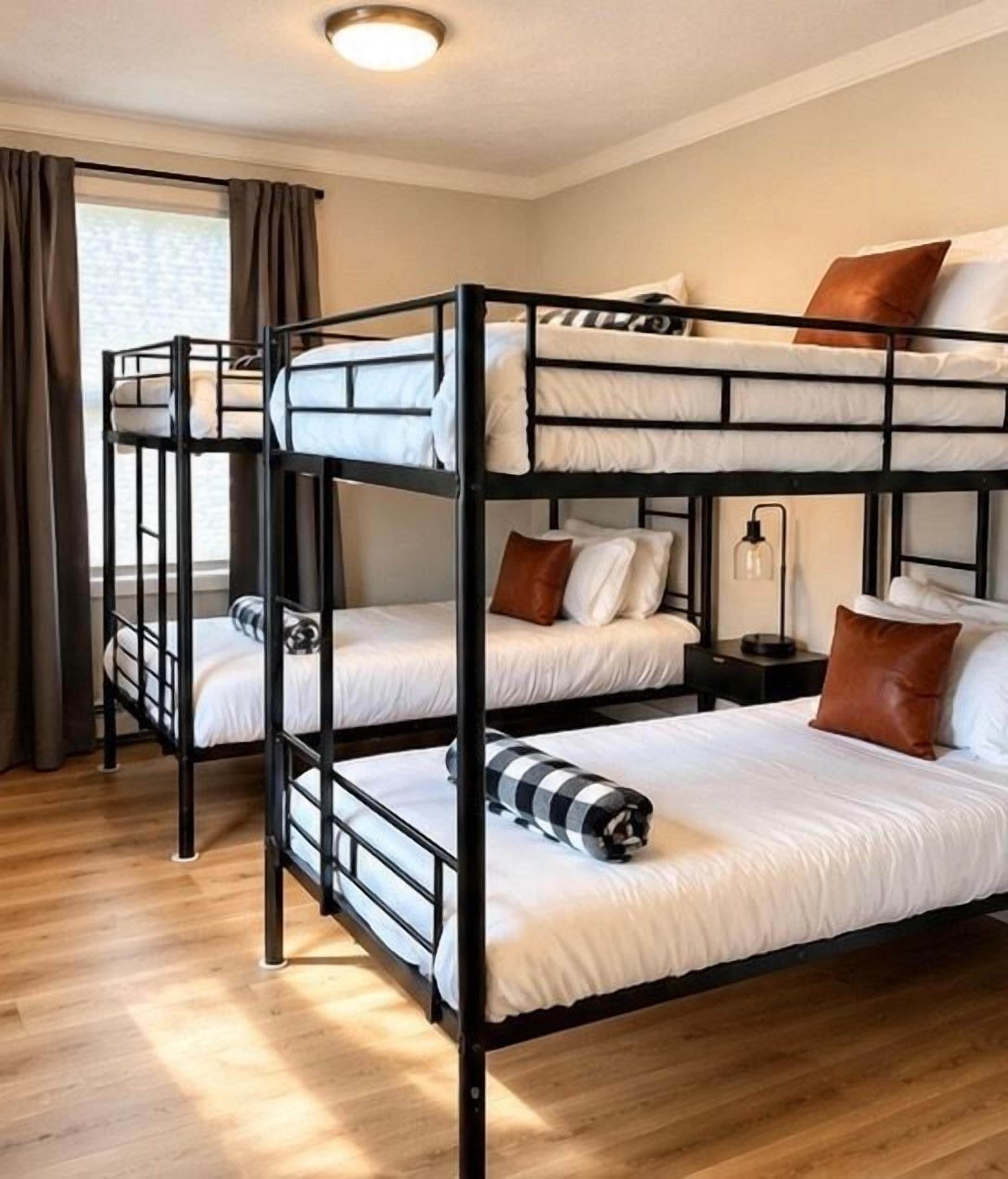 A bunk bed room with a wooden floor, a window with curtains, and a bedside table with a lamp.
