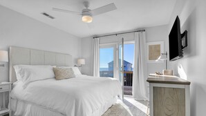 A bedroom with a large bed, a nightstand, a dresser, a ceiling fan, and a view of the outside. - The Shady Lady: Spacious Ocean View Home with Open Floorplan (Oak Island)