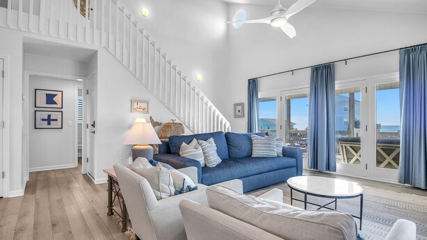 A spacious living room with a blue sofa, white armchairs, and a round coffee table. A staircase with white railings leads to an upper level. - The Shady Lady: Spacious Ocean View Home with Open Floorplan (Oak Island)