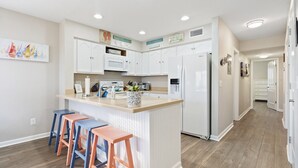 A modern kitchen with white cabinets, a central island with stools, a refrigerator, and a microwave. - Oceanfront Condo w/Views, Pool + Parking (Surf City)