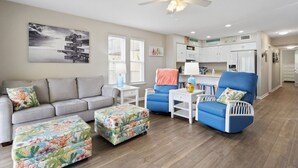 A living room with a grey sofa, blue armchairs, and colorful ottomans. A ceiling fan and a painting are visible on the wall. - Oceanfront Condo w/Views, Pool + Parking (Surf City)
