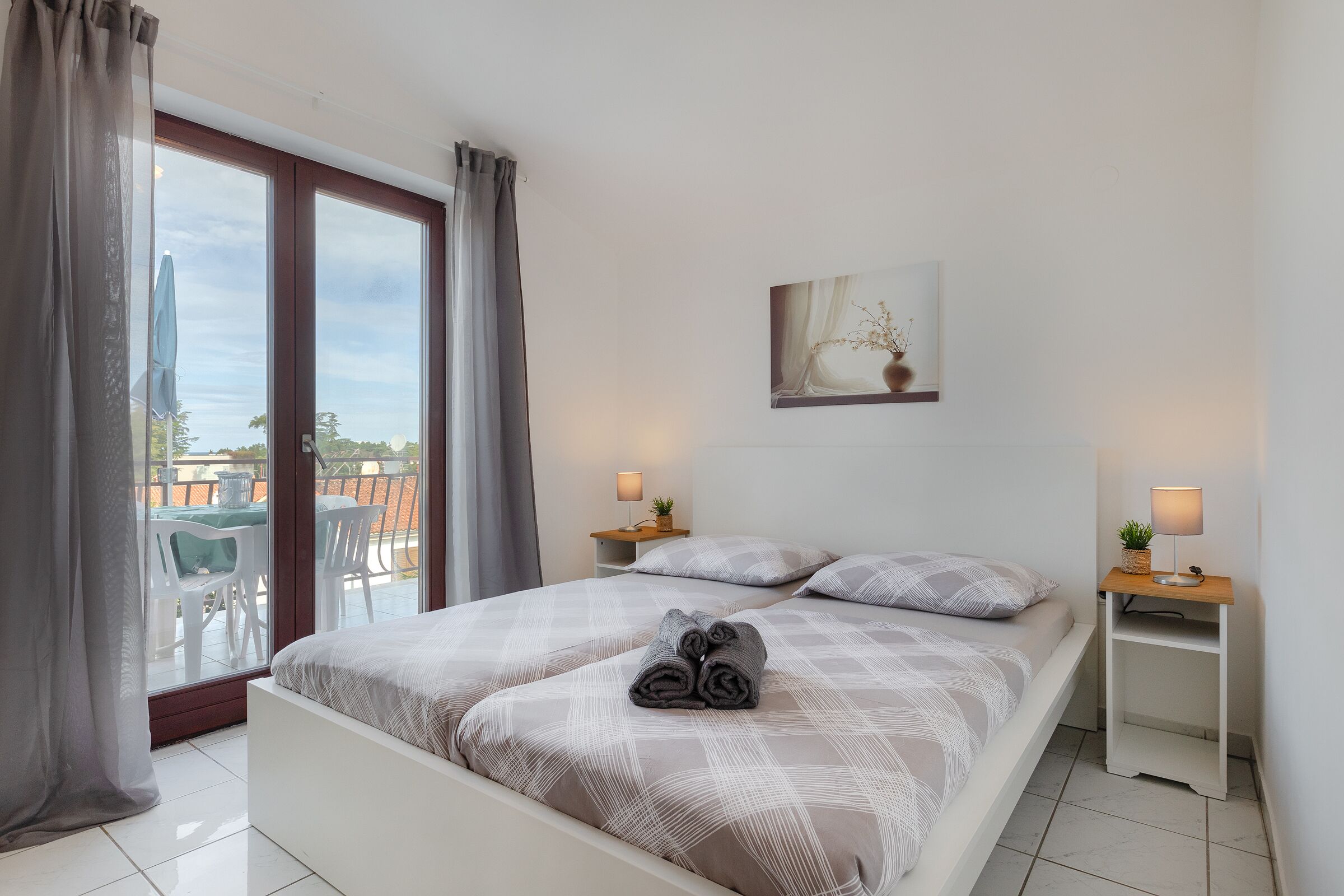 A bedroom with a bed, two bedside tables with lamps, a window with curtains, and a painting on the wall.