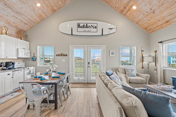 A spacious living area with a dining table, a sofa, and a kitchen with white cabinets and a wooden ceiling.