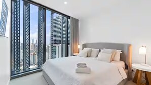 A modern hotel room with a large bed, a bedside table, and a view of the cityscape through floor-to-ceiling windows. - Signature 2B2B Stunning Chic Guest Fav +OceanView (Broadbeach)