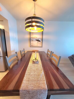 A dining area with a long wooden table, six chairs, a woven runner, and a hanging light fixture with a picture above.