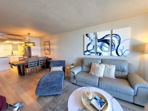 A modern living room with a grey sofa, a blue armchair, and a dining area with a wooden table and chairs. There is a large abstract painting on the wall and a ceiling fan above the dining table.