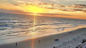 Ein Strand bei Sonnenuntergang, die Sonne tief am Horizont, sanfte Wellen am Ufer und einige Personen sowie ein Auto am Strand.