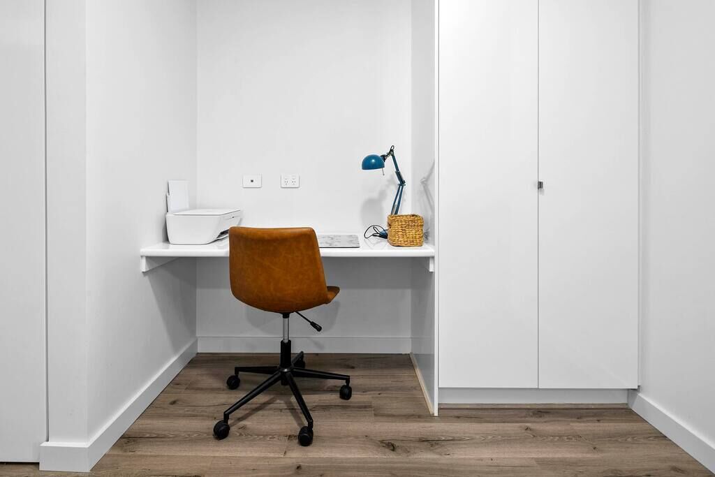 A small, clean workspace with a wooden floor, a brown office chair, a white desk, a blue desk lamp, and a wicker basket.