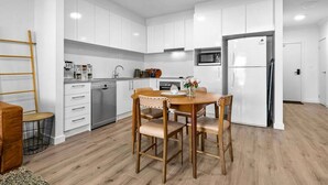 Eine moderne Küche mit weißen Schränken, einem Edelstahl-Geschirrspüler, einer Mikrowelle und einem Kühlschrank. Ein Holztisch mit vier Stühlen steht daneben.