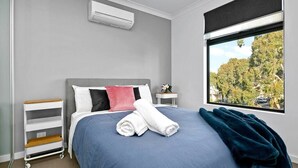 Ein Schlafzimmer mit einem Bett, einem Fenster mit Blick auf Bäume, einer Klimaanlage, einem Nachttisch und einem kleinen Regal.